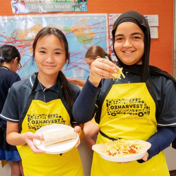 Two schoolgirls in yellow OzHarvest aprons hold plates of wraps and salad in a classroom