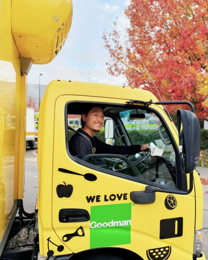 Smiling driver at the wheel of a yellow OzHarvest truck with autumn trees and 'We Love Goodman' decal