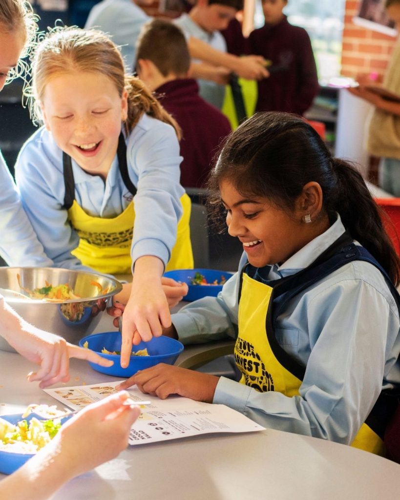 Primary school children in yellow OzHarvest aprons laughing while preparing food at a FEAST cooking class