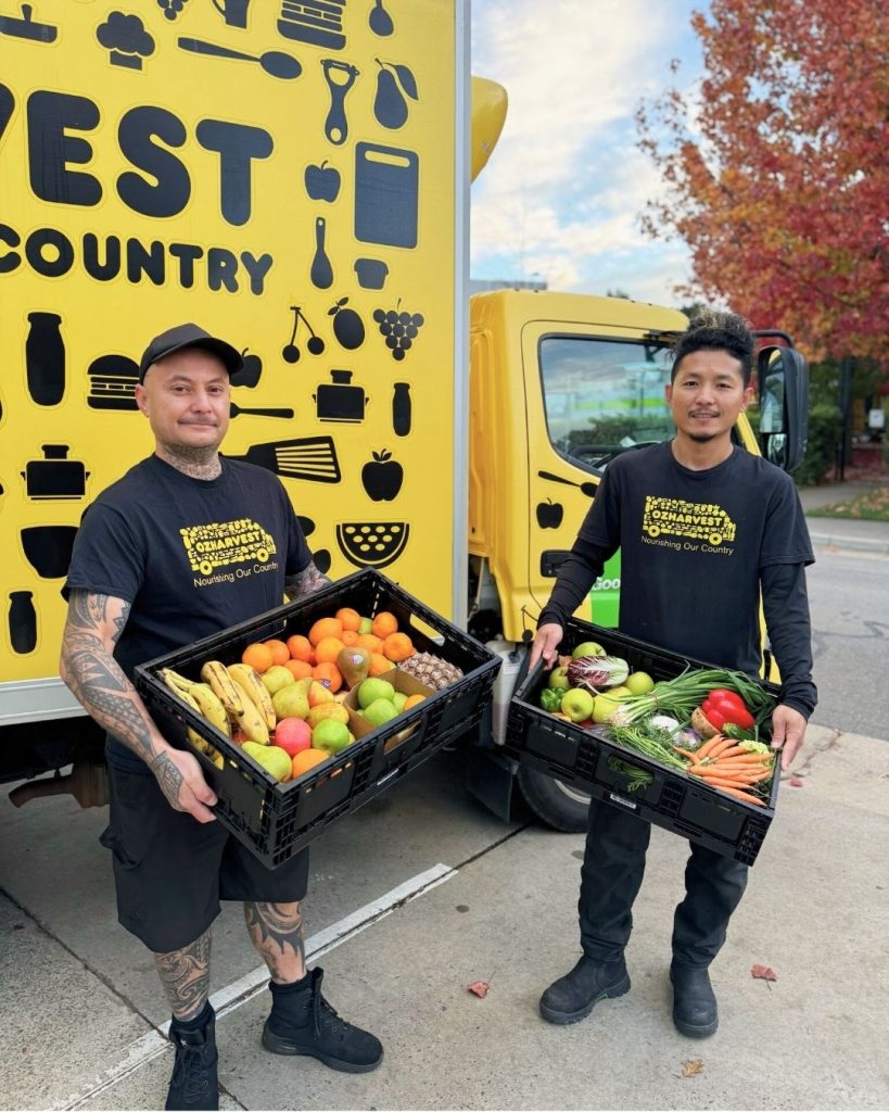 Two OzHarvest volunteers in branded shirts hold crates of rescued fruit and vegetables by a yellow truck