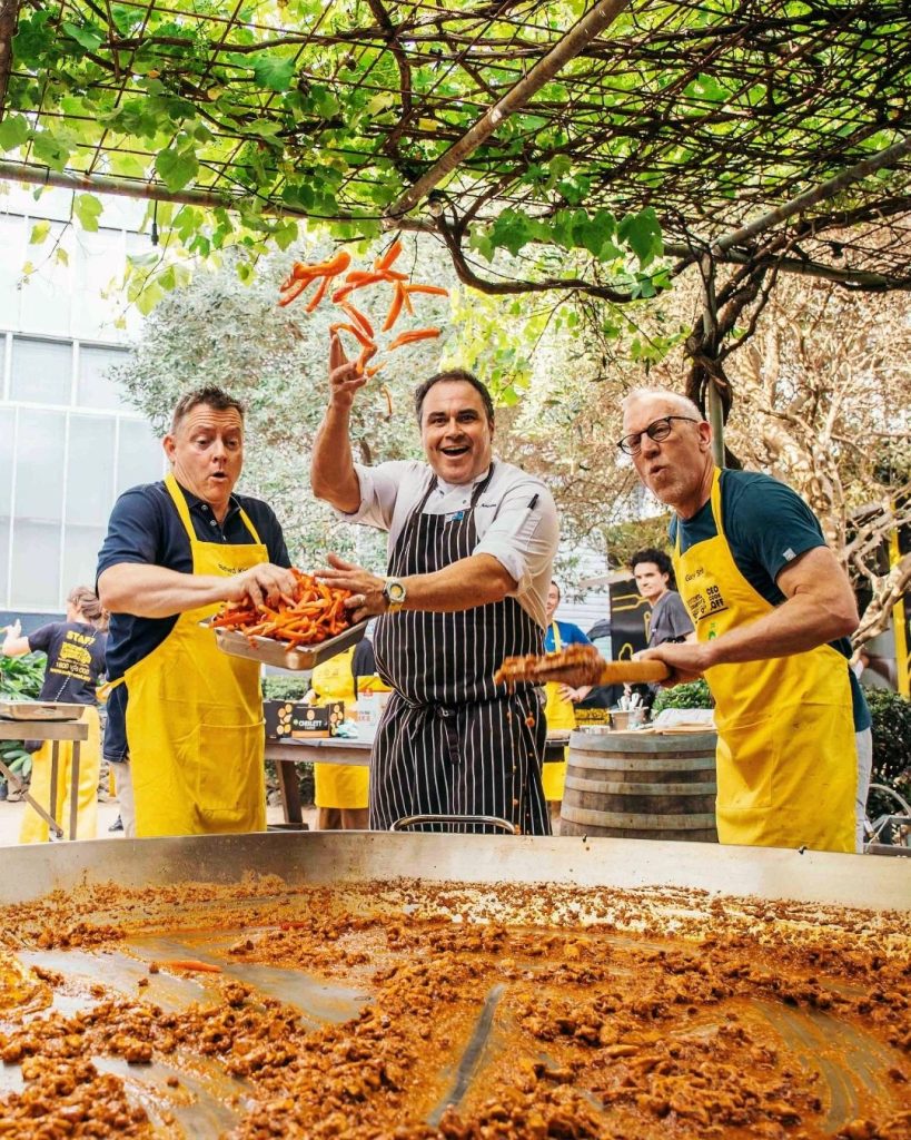 Three chefs toss chillies over a giant paella pan at an OzHarvest outdoor cook-up