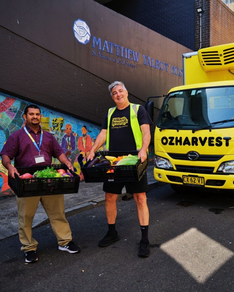 Two OzHarvest volunteers carry crates of fresh produce by a yellow truck outside Matthew Talbot Hostel