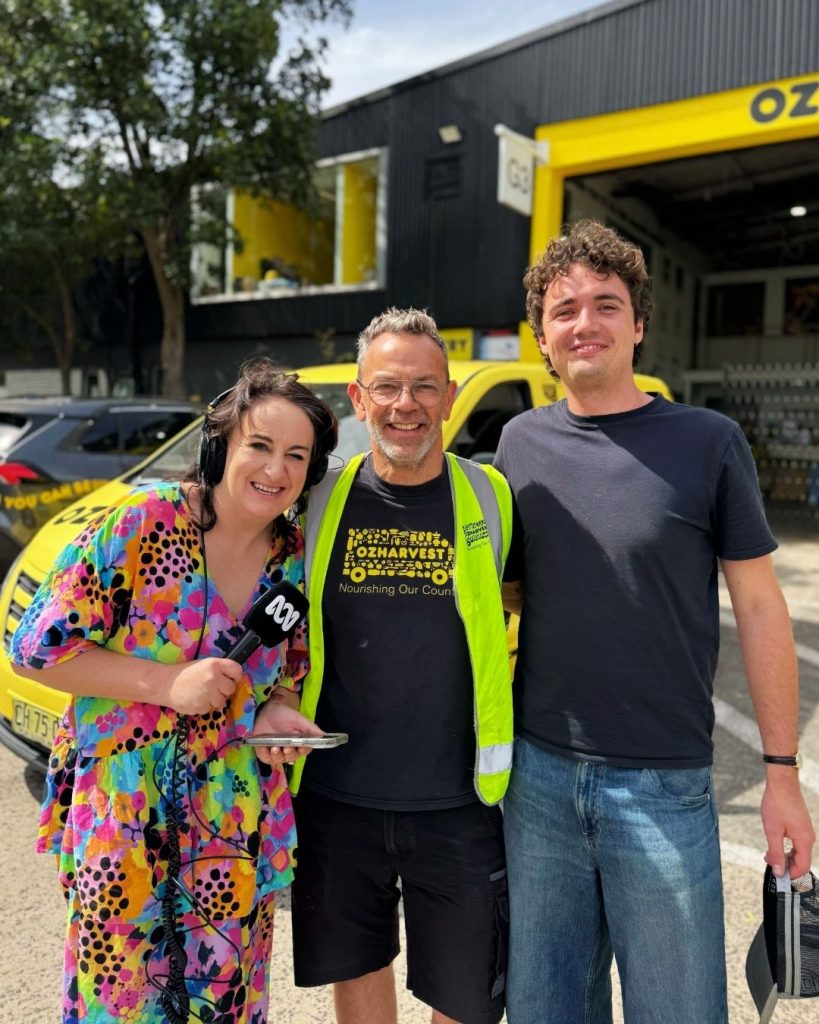 Radio reporter with ABC microphone interviews two smiling OzHarvest volunteers at the depot