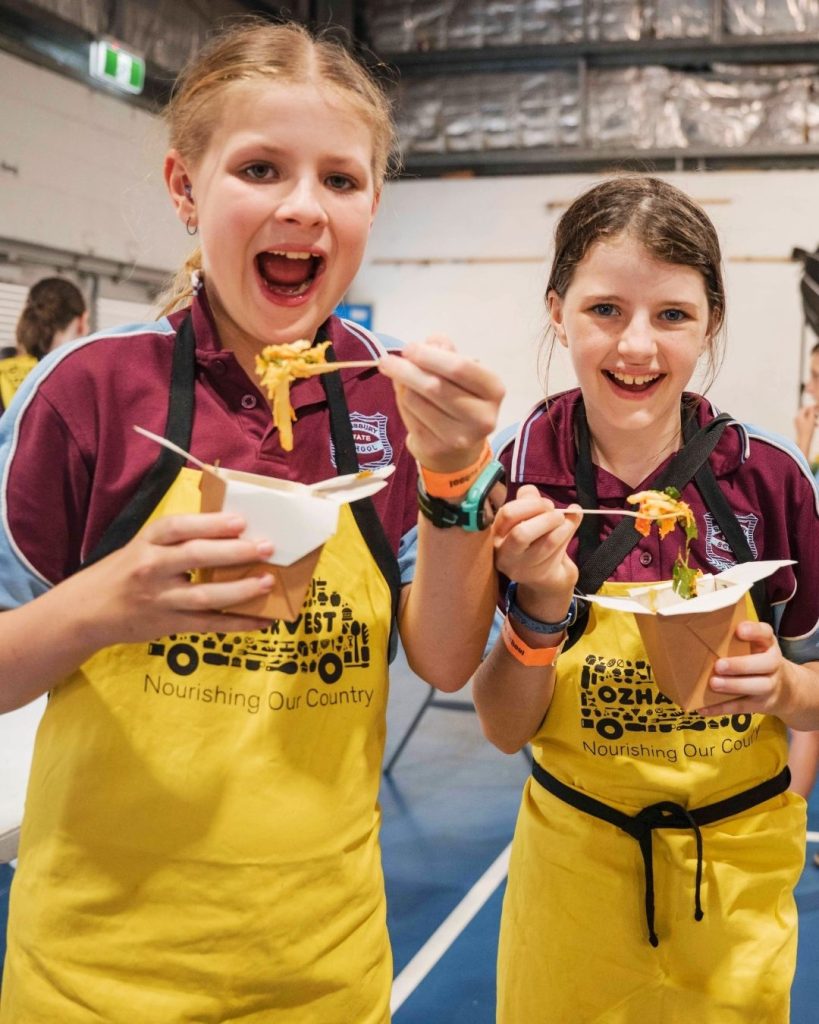 Two schoolgirls in maroon uniforms and yellow OzHarvest aprons laughing while eating noodles at a cooking event