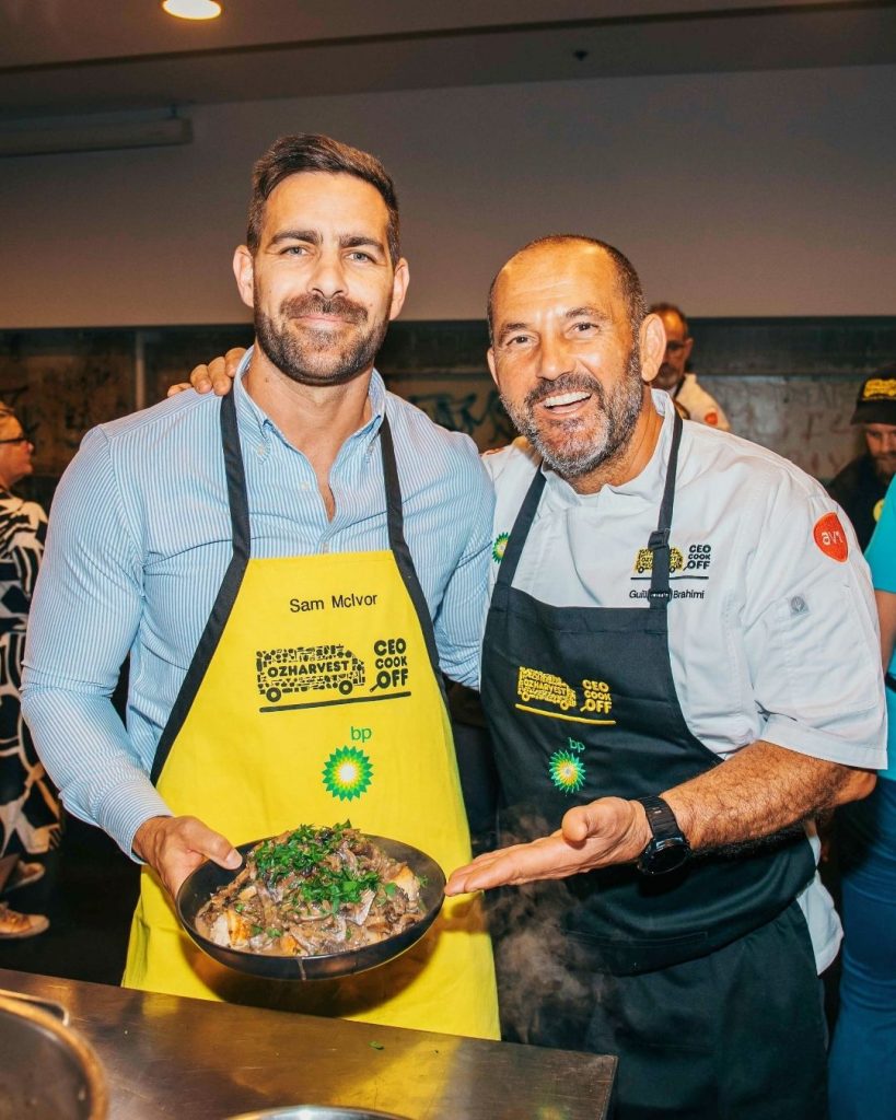 Two men in OzHarvest CEO CookOff aprons presenting a dish of cooked food at an event