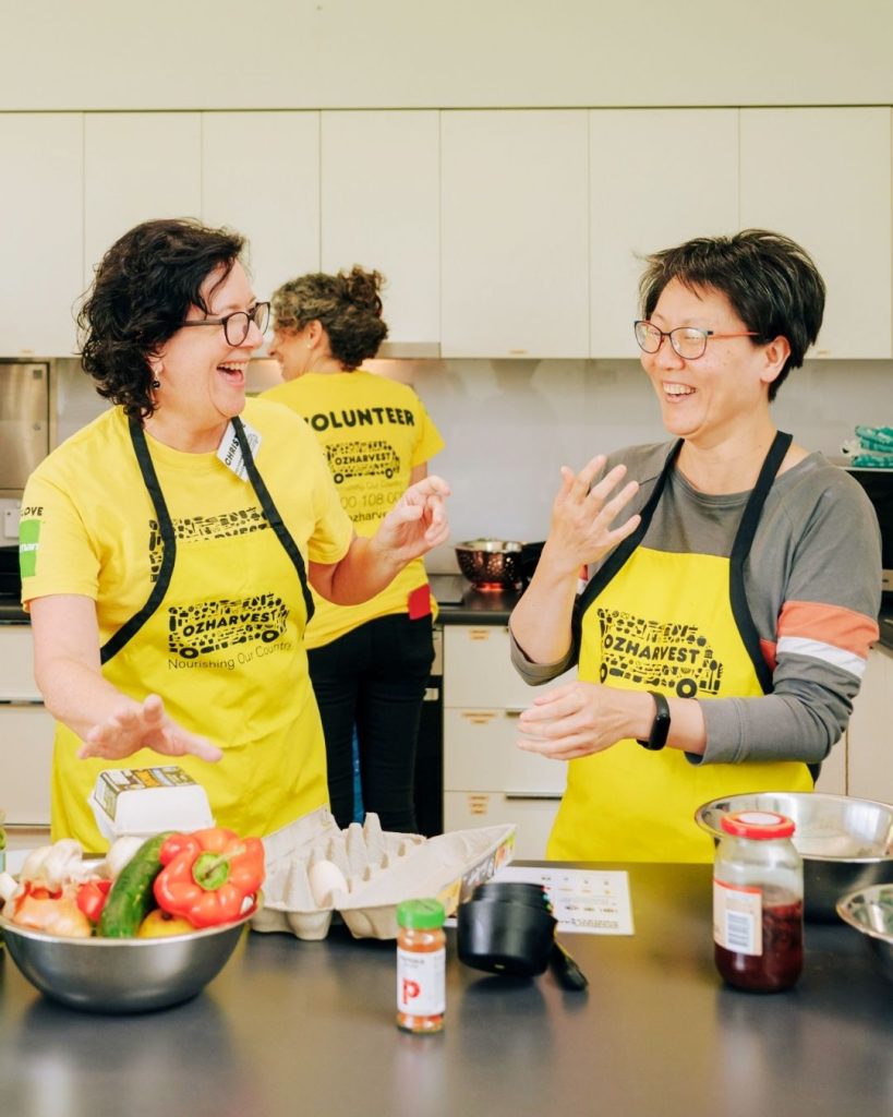 OzHarvest volunteers laughing while cooking together with fresh vegetables in a kitchen