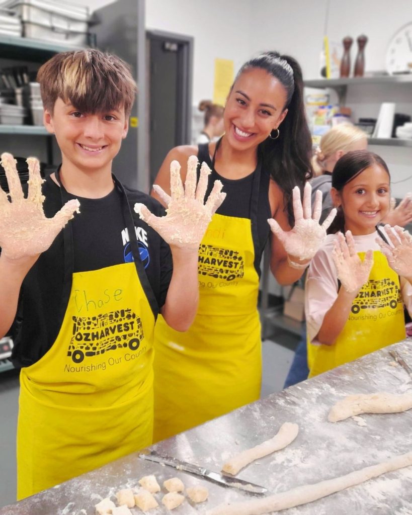Two children and a woman in OzHarvest aprons showing dough-covered hands while making gnocchi