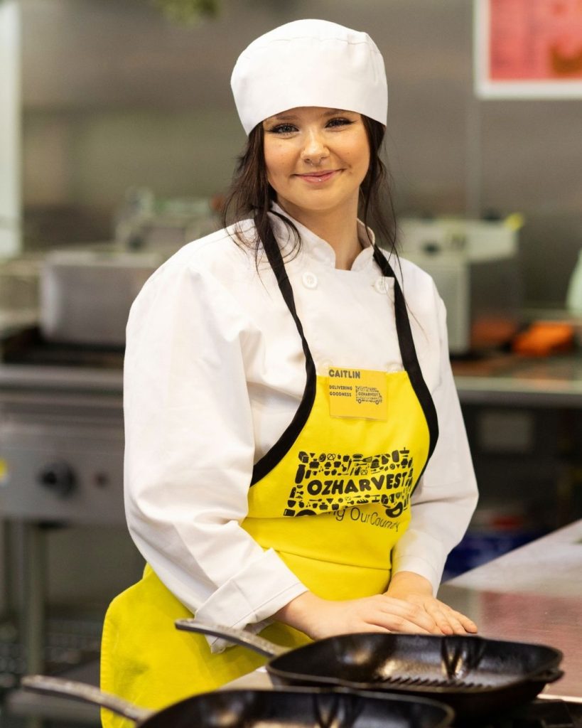 Young woman in chef whites and yellow OzHarvest apron smiling in a commercial kitchen