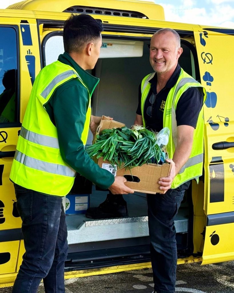 Two men in hi-vis vests loading a box of fresh greens into a yellow OzHarvest van