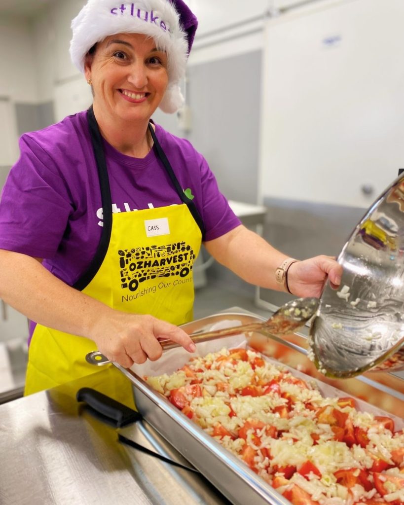 Smiling volunteer in OzHarvest apron and Santa hat serving rice and tomato salad from a tray