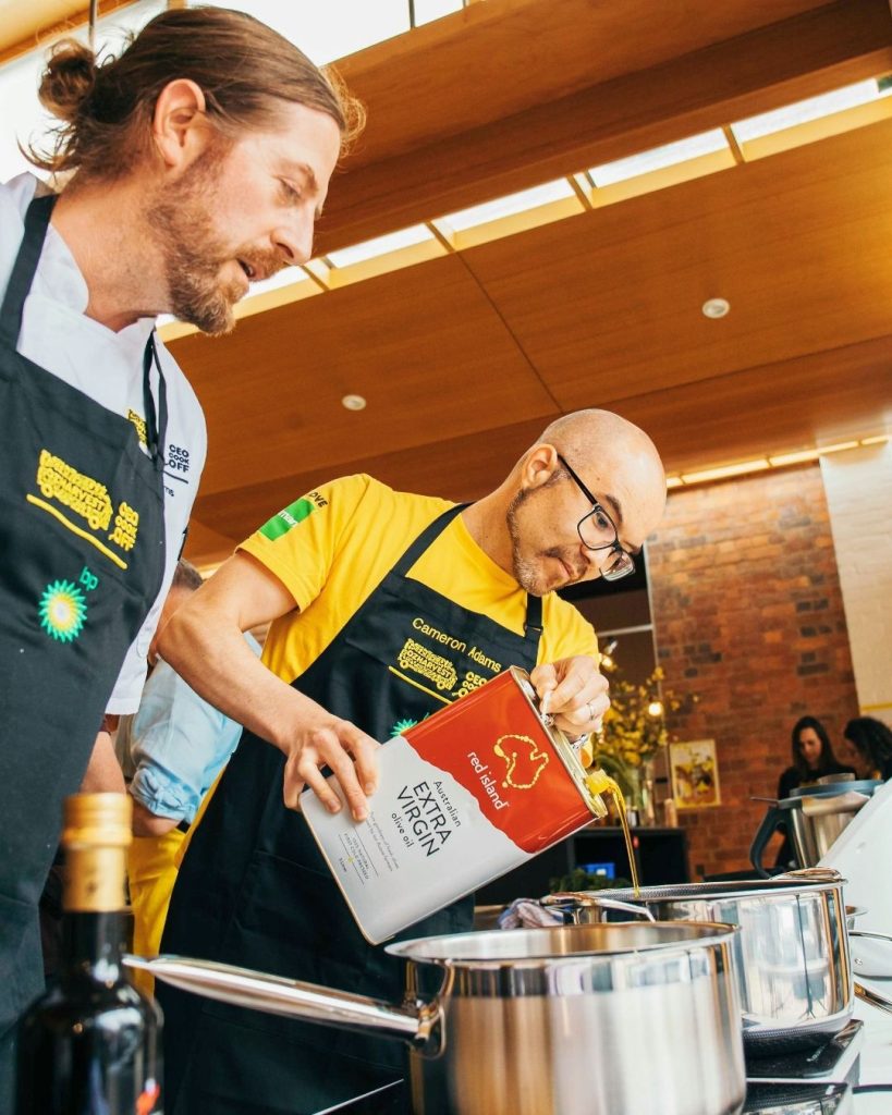 Chefs in OzHarvest CookOff aprons pouring olive oil into a pot at a cooking event