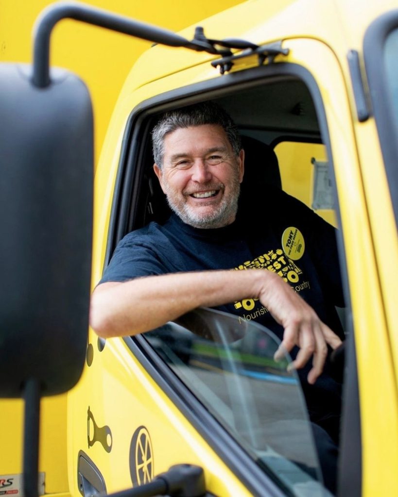 Smiling OzHarvest driver leaning out the window of a bright yellow truck