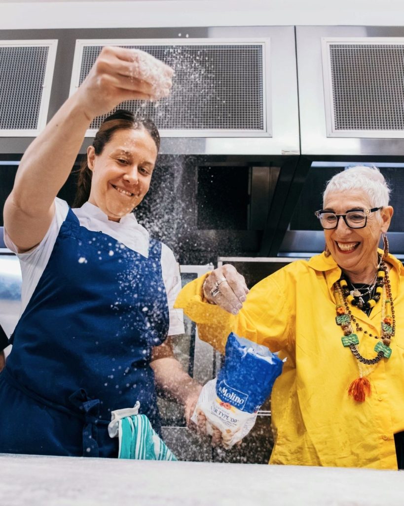 Two women laughing while dusting flour over a workbench in a commercial kitchen
