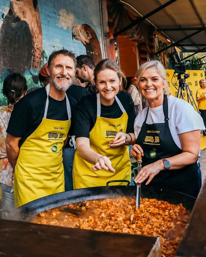Three smiling volunteers in yellow BP aprons cooking a giant paella at an OzHarvest Cooking for a Cause event