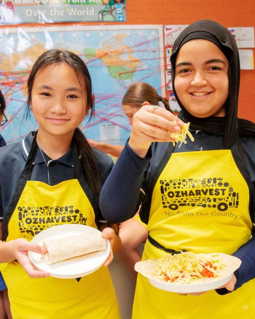 Two smiling schoolgirls in yellow OzHarvest aprons holding plates of wraps in a FEAST classroom