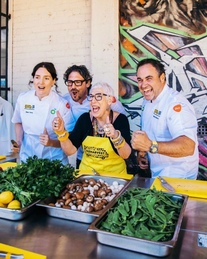 Chefs in white jackets and an OzHarvest apron cheering over trays of fresh vegetables at a cooking event