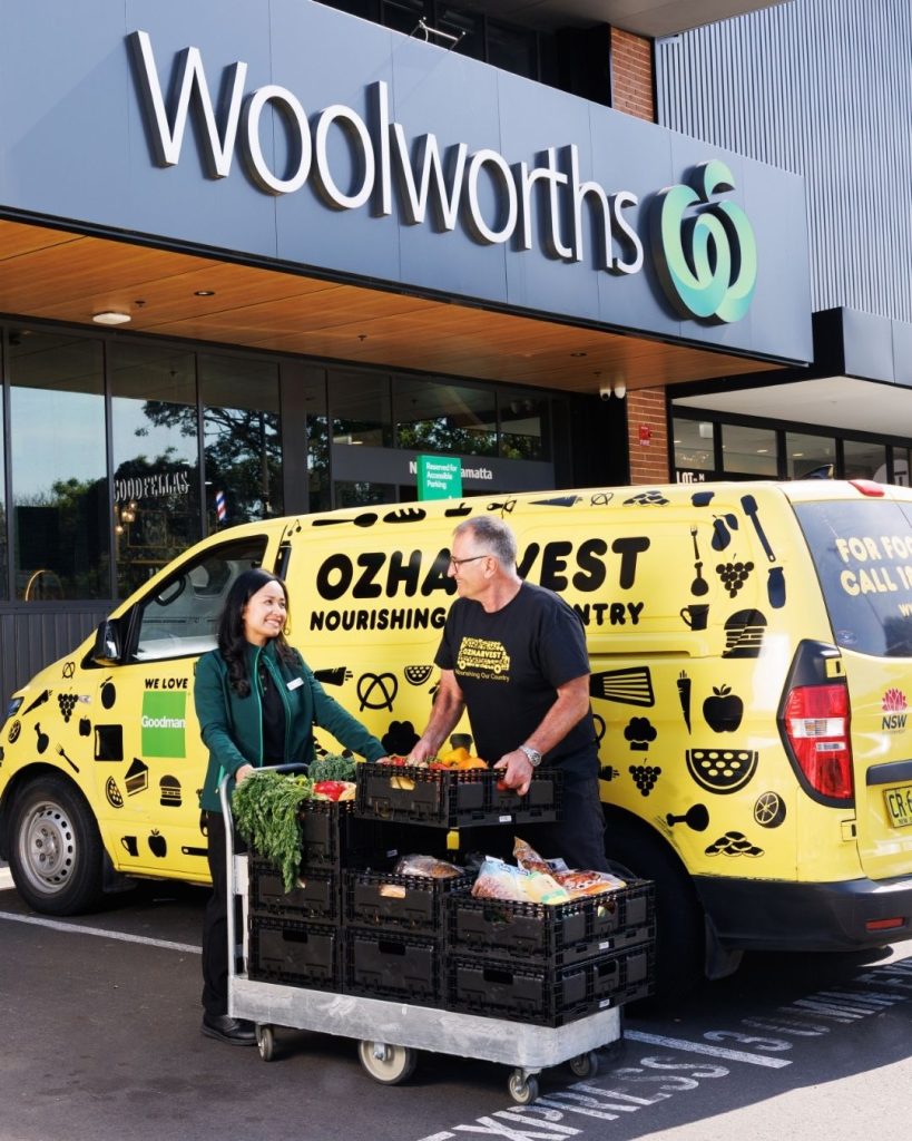 OzHarvest worker and a partner in a Goodman vest loading crates of rescued produce onto a trolley outside a Woolworths store