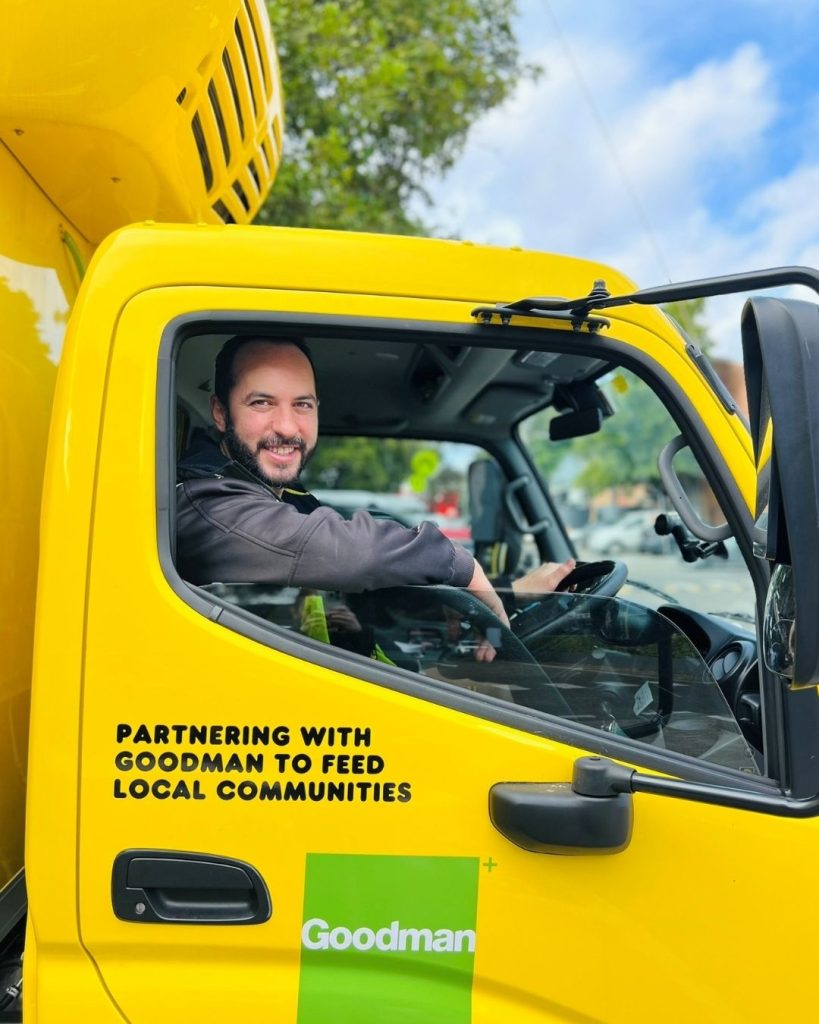Smiling driver in a yellow OzHarvest truck cab reading Partnering with Goodman to feed local communities