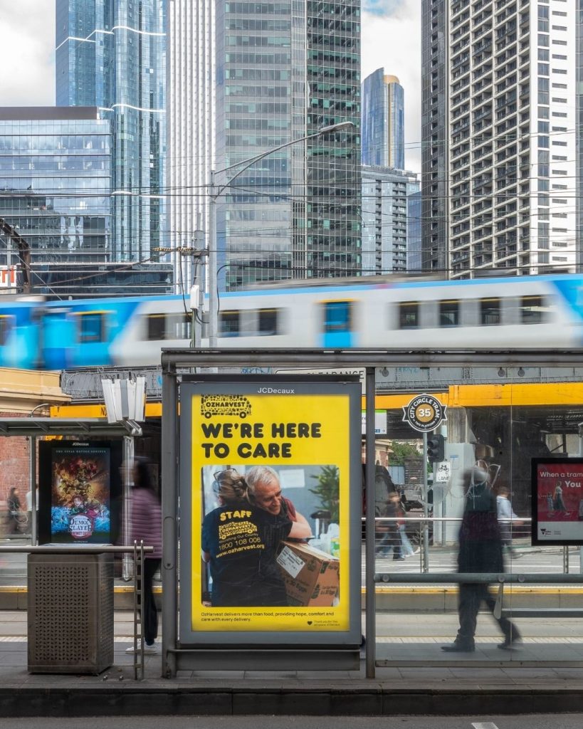 OzHarvest We're Here To Care advertisement on a city tram stop with a passing Melbourne tram