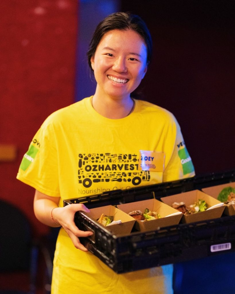 Smiling OzHarvest volunteer in a yellow shirt holding a crate of boxed meals at an event