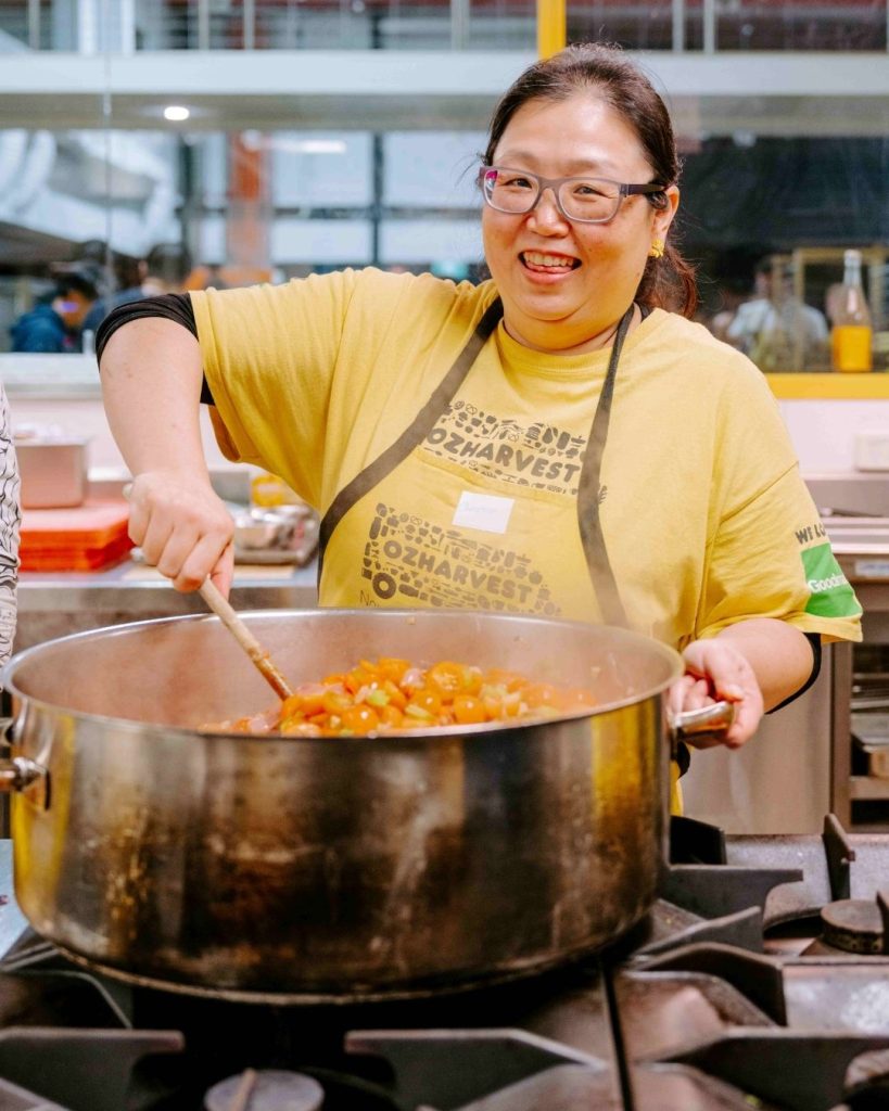Smiling OzHarvest volunteer stirring a large pot of tomato sauce on a stove in a community kitchen