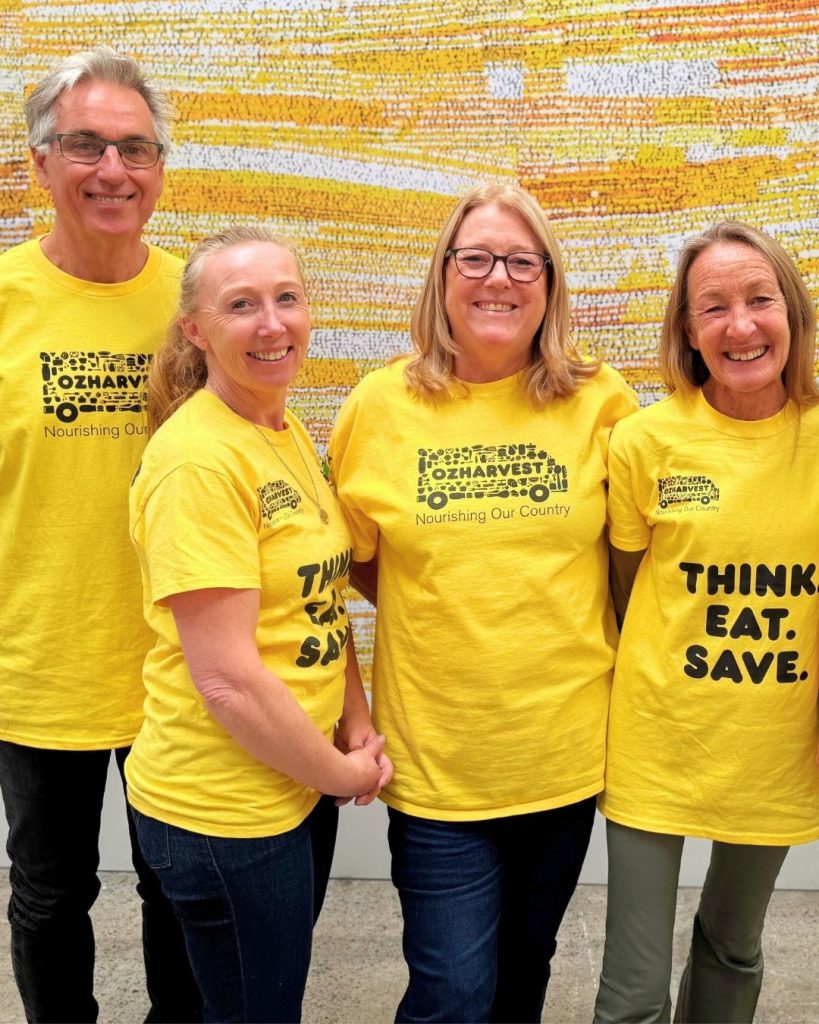 Four volunteers in yellow OzHarvest 'Think. Eat. Save.' shirts smile in front of a colourful mural