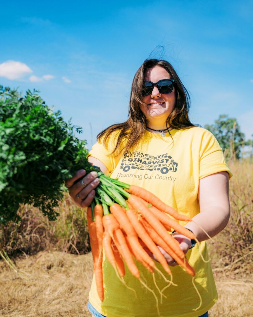 Smiling woman in a yellow OzHarvest t-shirt holding a freshly pulled bunch of carrots in a sunny field
