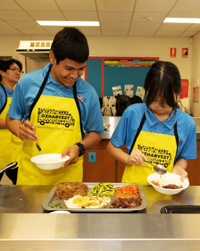 Two students in blue uniforms and yellow OzHarvest aprons serving vegetables into bowls in a school cooking class