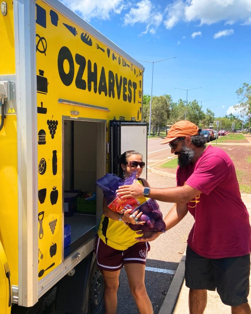 Man handing a mesh bag of onions to a smiling woman beside an open yellow OzHarvest truck on a sunny street