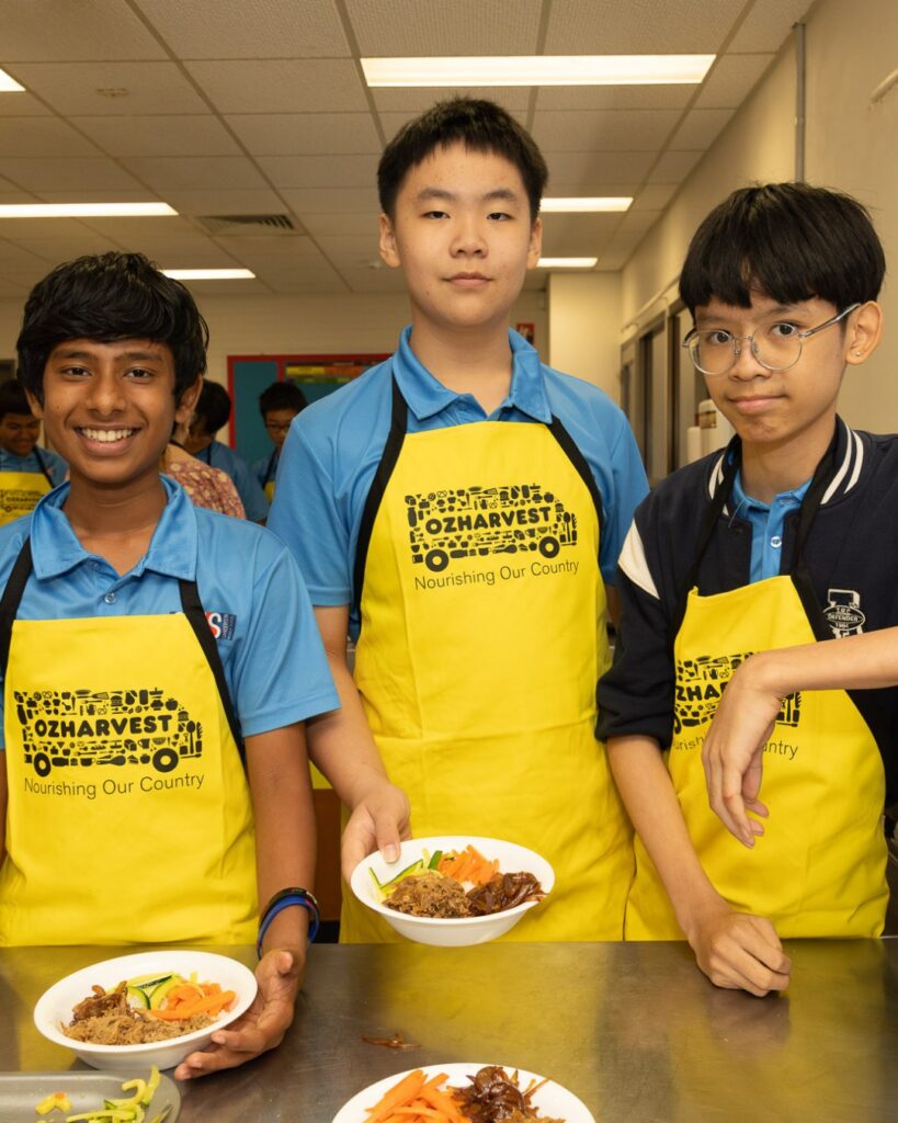 Three students in yellow OzHarvest aprons and blue uniforms at a kitchen bench, one holding a bowl of food